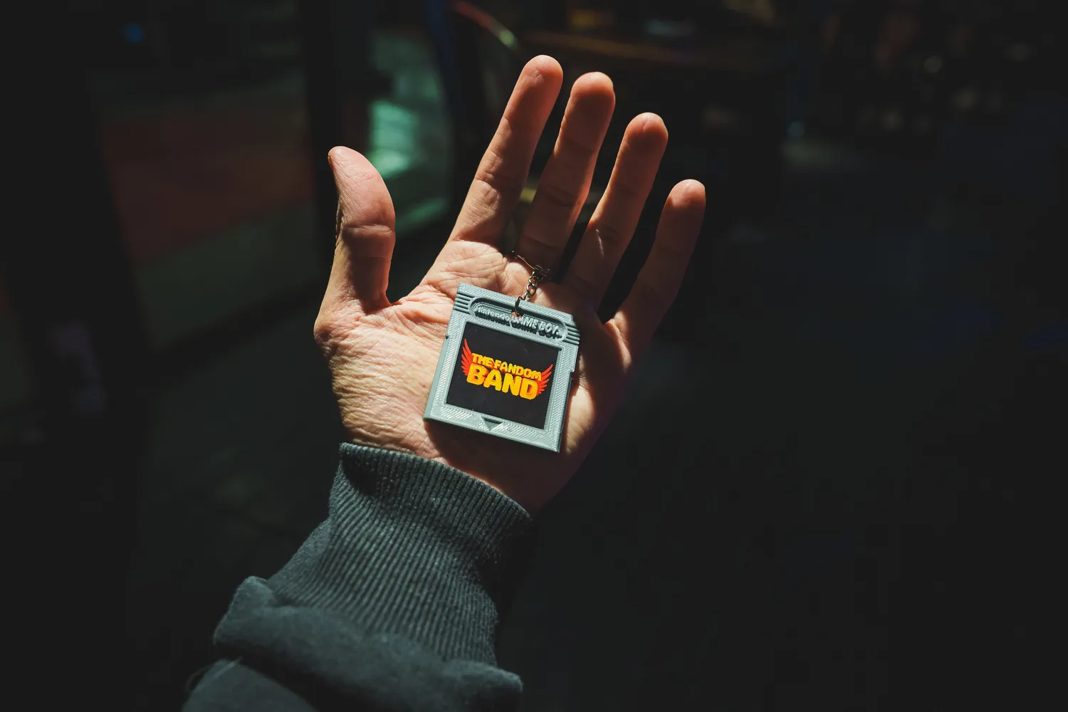 Una mano sostiene un llavero con forma de cartucho de Game Boy con el logo de The Fandom Band